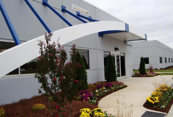 Front entrance of Turbomeca TMM Manufacturing Facility. Awning supported by blue structures. Floral landscaping.